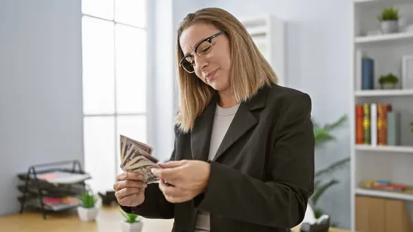 Attractive young blonde business woman in glasses brimming with happiness as she counts her many dollars at the office workstation