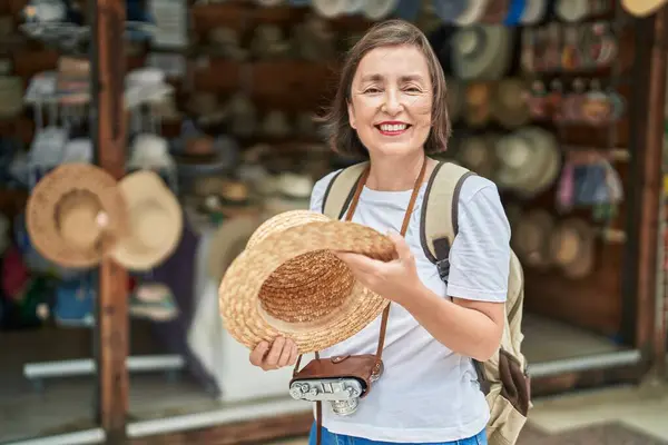Middle age woman tourist smiling confident holding hat at street market