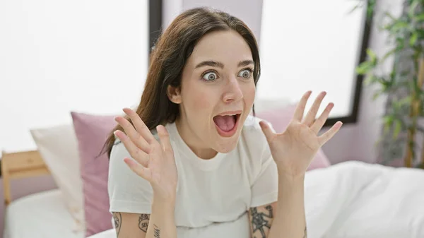 A surprised young caucasian woman expressing excitement indoors, with a tattoo visible and a bedroom setting.