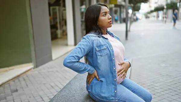 Tired young pregnant woman, touching her belly, sits in contemplation on an urban street bench, relaxed, balancing the emotion of expectant motherhood and looming maternity ache.