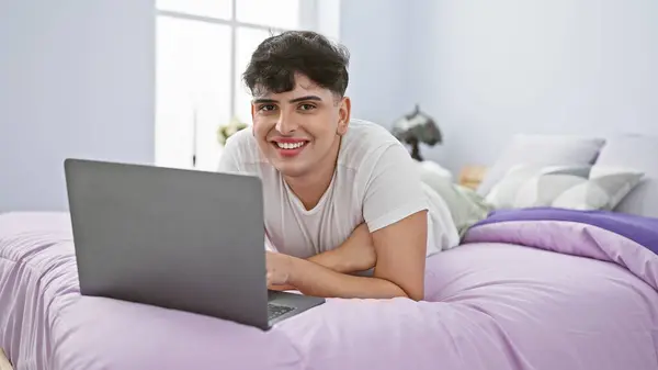 A smiling man lying on a purple bed using a laptop in a bright bedroom