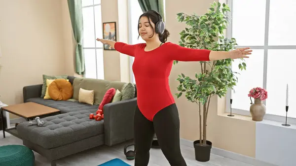 A young hispanic woman enjoys exercise with headphones in a well-lit living room, exuding wellness and happiness.
