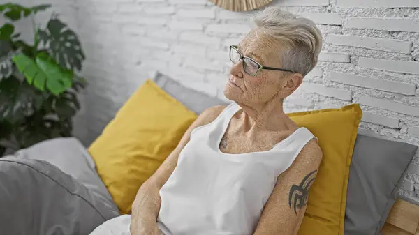 Ciddi görünümlü, gri saçlı yaşlı kadın rahat bir şekilde rahat yatağında dinleniyor, derin düşünceler içinde, sade yatak odasının sıcaklığında, soluk bir sabah ışığı içeri sızıyor.