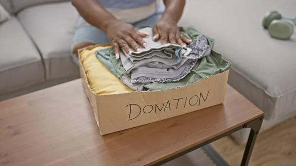 Warm-hearted african american woman putting folded clothes into a cardboard box for charity donation, comfortably sitting on her living room sofa, turning chores into support at home