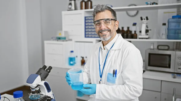 Cheerful young grey-haired hispanic man scientist confidently measuring liquid in test tube, working indoors at a busy laboratory, smiling under safety glasses and gloves