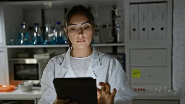 Hiszpanka Białym Fartuchu Laboratoryjnym Badająca Tabletkę Słabo Oświetlonym Pomieszczeniu Laboratoryjnym — Zdjęcie stockowe