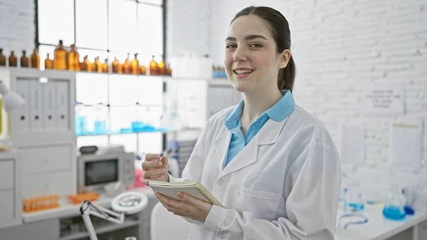 Confident young woman in a lab coat holding a notebook inside a modern laboratory with equipment and glassware.