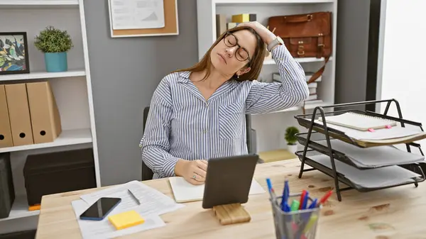 A tired young woman with glasses stretching at her organized office desk.