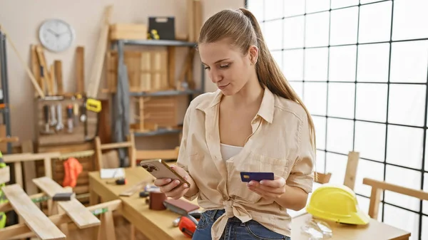 A young woman in casual attire holding a credit card and using a smartphone in a well-lit carpentry workshop