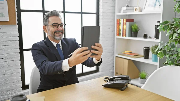 Handsome, grey-haired, young hispanic business man takes a radiant smiling selfie with touchpad at his office, rocking success in suit and tie.