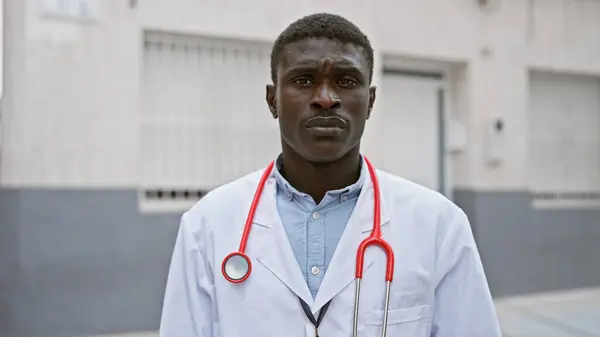 Confident african man in a lab coat with a stethoscope standing outside in a city.