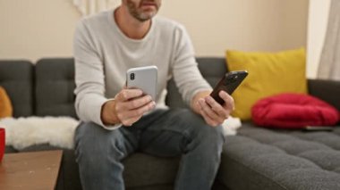 Orta yaşlı sakallı adam akıllı telefonları evde rahat kullanıyor, çağdaş yaşam tarzı ve teknoloji entegrasyonunu yansıtıyor..