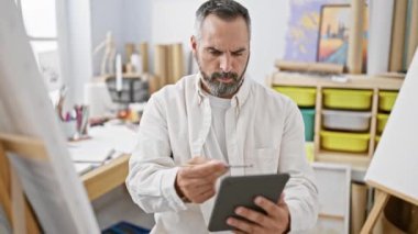 Sanat stüdyosunda tablet kullanan sakallı ve gri saçlı olgun bir sanatçı.