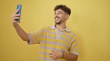 Neşeli genç Arap adam kendinden emin bir video çağrısına sevinçle dalıyor, sarı izole edilmiş parlak bir arka plana karşı gelişigüzel duruyor..