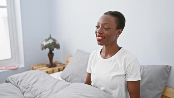 喜びを受け入れる楽しいアフリカ系アメリカ人女性 彼女の居心地の良い寝室でカメラで抱擁と笑顔のために広い腕でベッドに横たわる — ストック動画
