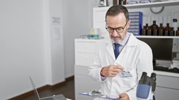一个白发苍苍 自信的中年男人 穿着实验室外套面带微笑 一丝不苟地在一个忙得不可开交的实验室的剪贴板上记笔记 — 图库视频影像