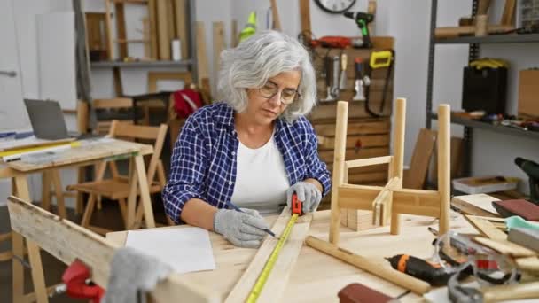 成熟した灰色の髪の女性は設備の整った大工のワークショップで木材を測定します — ストック動画
