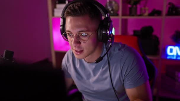 メガネとヘッドフォンを着用したヒスパニックな男性が 鮮やかな紫色のライトで照らされた部屋でゲームのセットアップに座ります — ストック動画