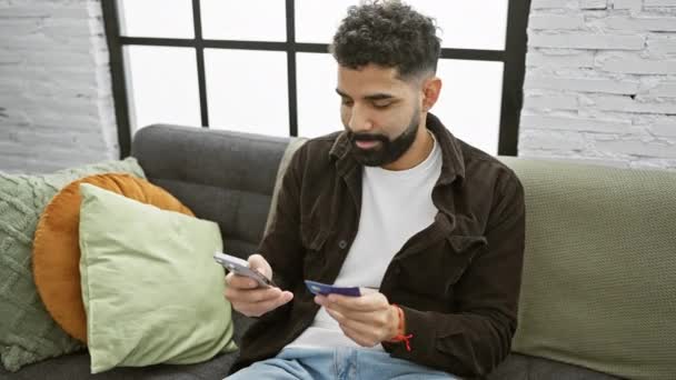 一个满脸胡须的男人把信用卡放在室内 舒服地坐在一张绿色的沙发上 在一间现代化的客厅里用智能手机浏览 — 图库视频影像