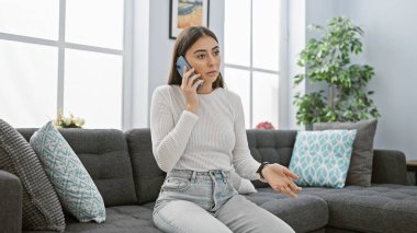 Modern bir oturma odasında akıllı telefondan konuşan İspanyol bir kadın, gündelik, rahat ve konuşmayla meşgul görünüyor..