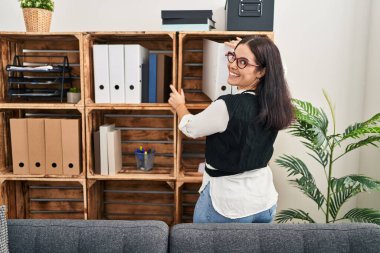 Young beautiful hispanic woman business worker holding documents of shelving at office