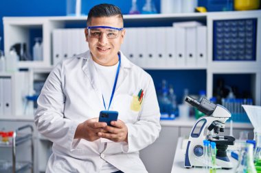 Young latin man scientist smiling confident using smartphone at laboratory