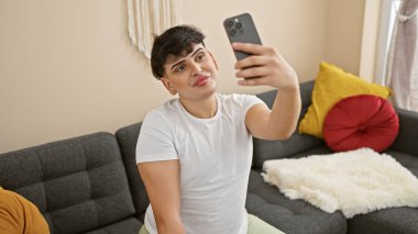 Genç bir adam cep telefonuyla selfie çekiyor, modern oturma odasındaki renkli minderlerle bir koltukta oturuyor..