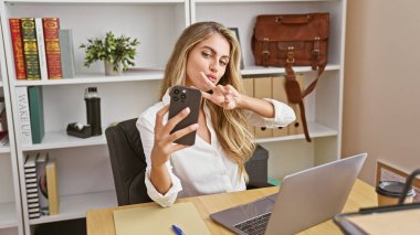 Parlak sarışın kadın işçi, kendine güven ve olumluluk saçan başarılı iş gününün tadını çıkarıyor. Bilgisayarda çalışırken şık ofis içi akıllı telefonuyla özçekim fotoğrafı çekiyor..