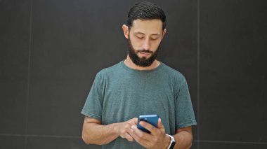 Genç İspanyol adam izole edilmiş siyah arka plan üzerinde akıllı telefon ve kulaklık kullanıyor.