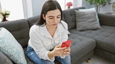 Modern bir oturma odasında oturan genç İspanyol kadın akıllı bir telefona odaklanmış, günlük ev teknolojisi kullanımını resmediyor..