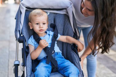 Anne ve oğlu sokakta bebek arabasında otururken kendinden emin bir şekilde gülümsüyor.