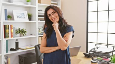 Modern ofis içinde duran, kendinden emin, orta yaşlı, gözlüklü bir kadın..