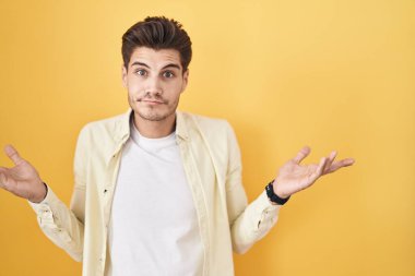 Young hispanic man standing over yellow background clueless and confused expression with arms and hands raised. doubt concept. 