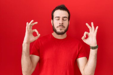 Latin kökenli genç bir adam, günlük kırmızı tişört giyiyor rahatlamış ve gözleri kapalı gülümsüyor parmaklarıyla meditasyon hareketi yapıyor. yoga konsepti. 