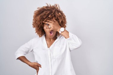 Beyaz arka planda duran kıvırcık saçlı genç İspanyol kadın. Şok olmuş bir şekilde yüzünü ve gözlerini kapıyor. Utanmış bir ifadeyle parmaklarının arasından bakıyor.. 