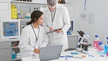 A woman and man, seemingly coworkers, analyze data together on a laptop in a well-equipped laboratory setting. clipart