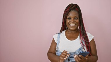 Kendine güvenen Afro-Amerikalı kadın akıllı telefondan sevinçle mesaj atıyor, pembe arka planda gülümsüyor.