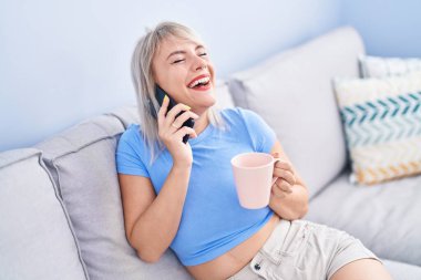 Young blonde woman talking on smartphone drinking coffee at home