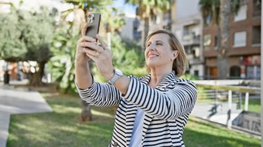 Olgun sarışın kadın güneşli bir parkta selfie çekiyor.