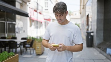 Young hispanic man using smartphone with serious expression at coffee shop terrace