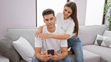 Bir kadın sıcak bir oturma odasında akıllı telefonuna bakan bir adamın omzuna şefkatle dokunur..