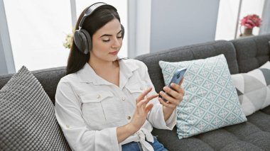 Hoş bir oturma odasında akıllı telefonunu karıştırırken kulaklıkla müzik dinlemekten hoşlanan çekici genç İspanyol kadın..