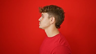 Havalı, genç İspanyol bir adam, güneşli ışıkta dikilip, izole edilmiş kırmızı duvar arkasında rahat bir yaşam tarzını resmediyor.