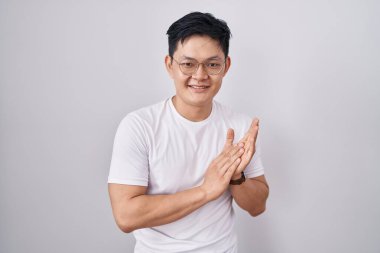 Genç Asyalı adam beyaz arka planda ayakta alkışlıyor ve alkışlıyor mutlu ve neşeli, gururla gülümsüyor. 