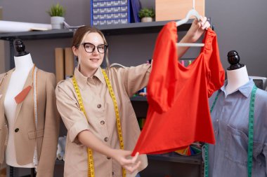 Young caucasian woman tailor smiling confident holding t shirt at atelier