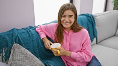 Genç ve güzel İspanyol kadın evdeki kanepede oturup kahve içiyor.