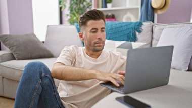 Çekici İspanyol genç adam, konsantre ve ciddi, rahat dairesinde yerde oturuyor, bilgisayarında çalışıyor, teknolojinin büyüleyici dünyasına dalmış..