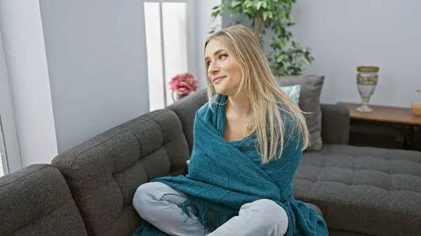 Chilled, young blonde woman snug in a blanket, yearning for warmth, looking through her home window