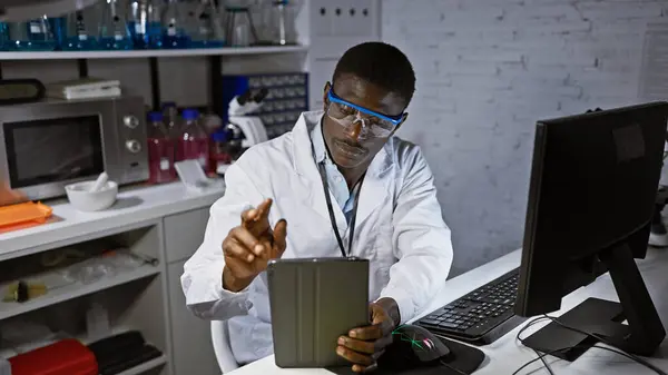 An african male scientist analyzes data on a tablet in a modern laboratory setting with computer and safety goggles.