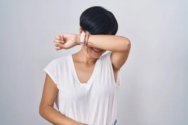 Young asian woman with short hair standing over isolated background covering eyes with arm smiling cheerful and funny. blind concept.